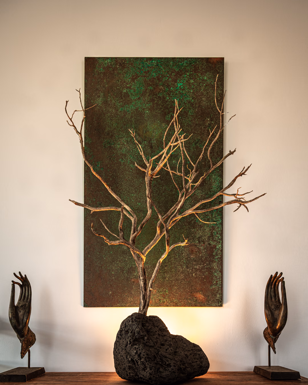 A pale dead branch seated in a block of lava rock and lit from behind, against a verdigris copper panel