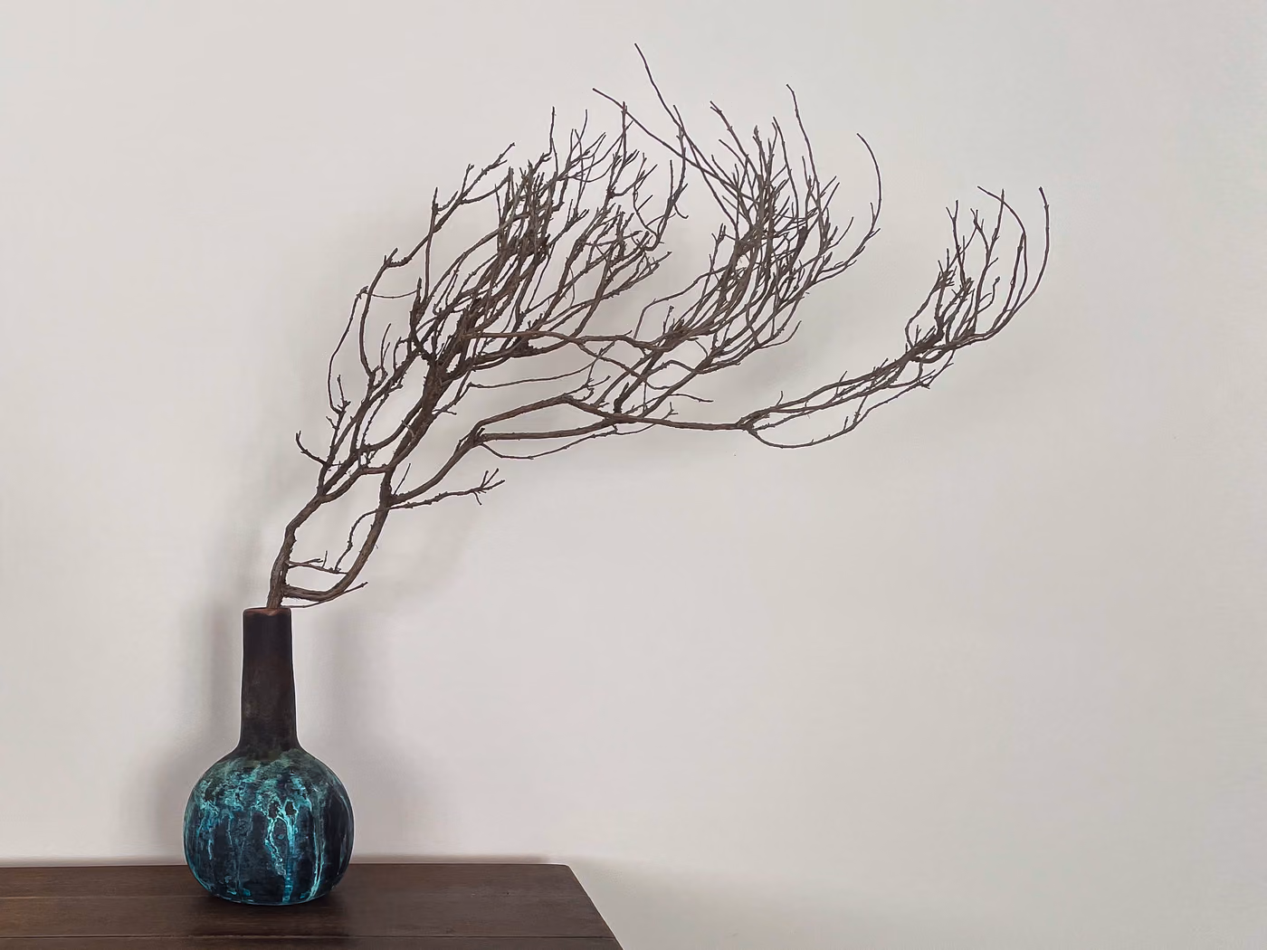 A dried rosemary branch sweeping out of a patinated blue-green vase on a dark wood surface against a plain pale wall